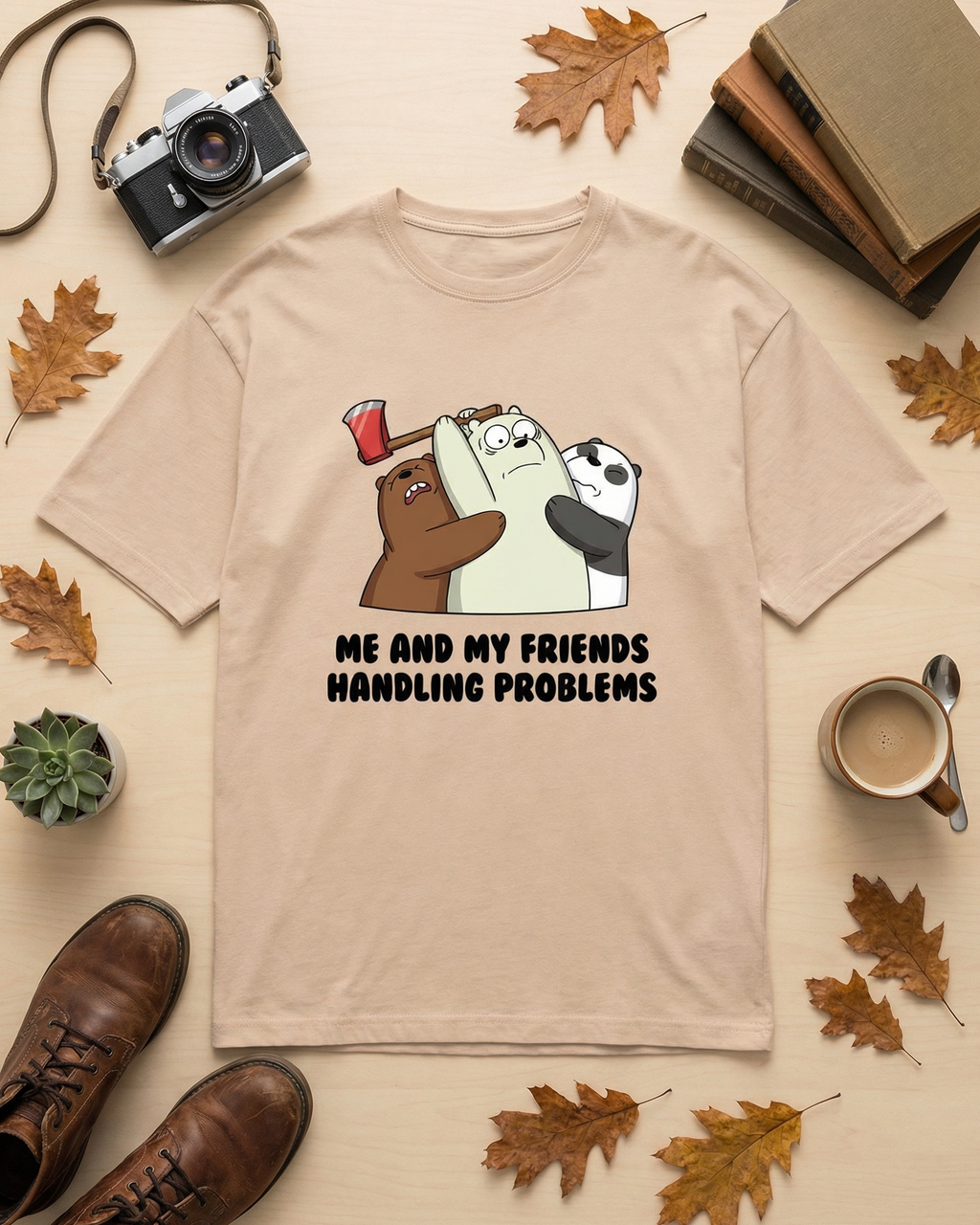 Beige t-shirt with cartoon design and text 'Me and my friends handling problems' on a surface with autumn leaves, books, and a camera.